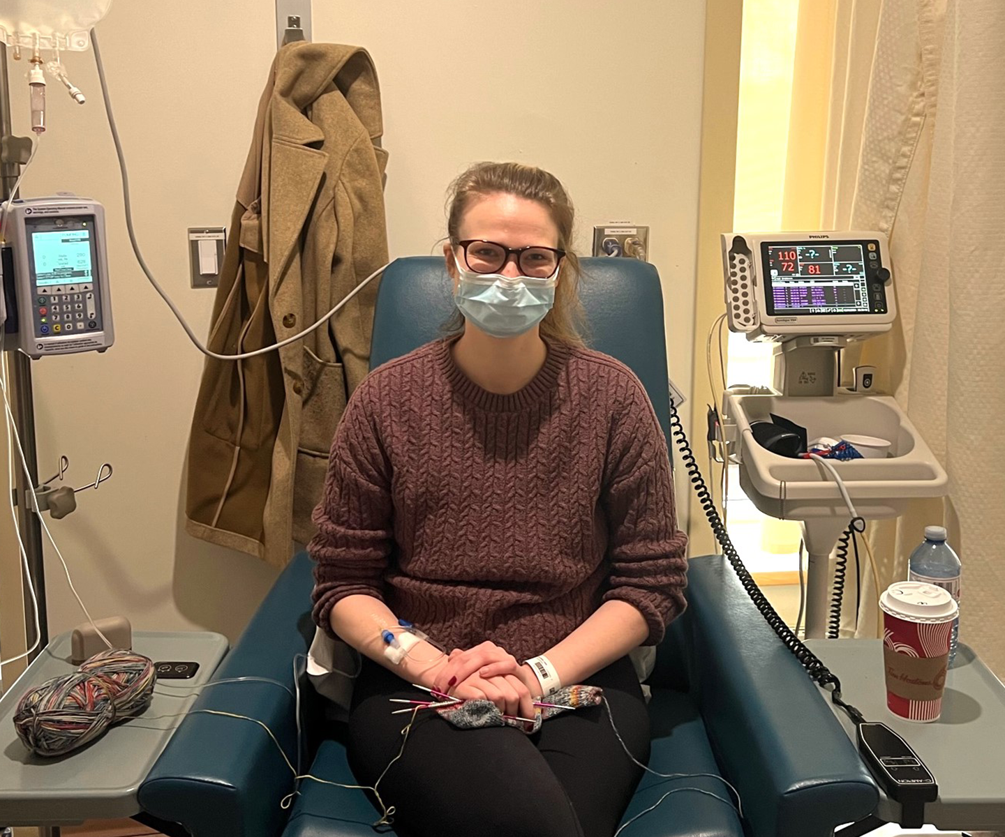 Masked person sits in blue hospital chair with intravenous tubes in one arm and a small knitting project in lap. A large bag of immunoglobulin being infused hangs above.  