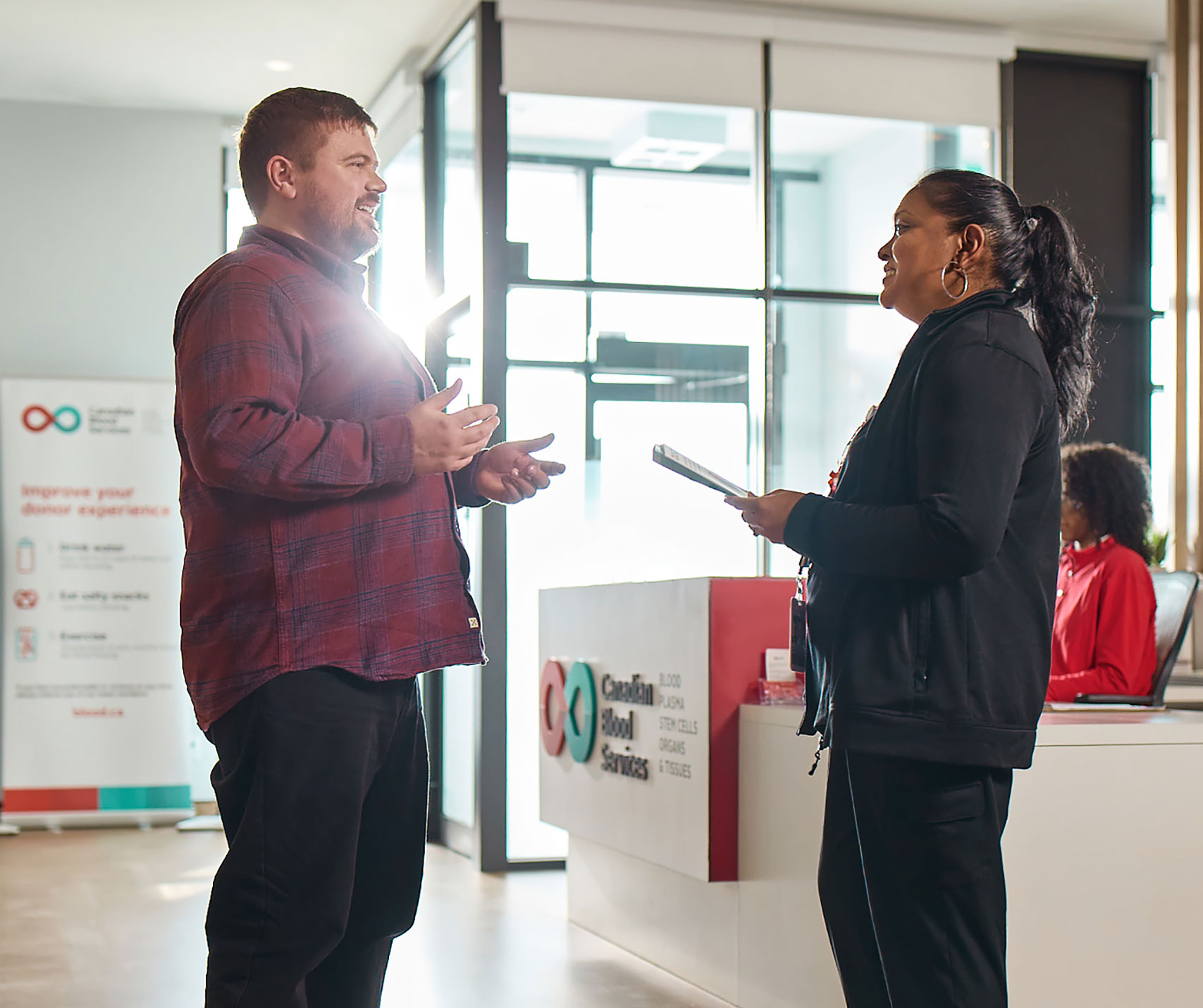 Ben, a blood donor, standing inside of a donor centre talking with a nurse. They are facing eachother while having a pleasant conversation in a bright, sun-filled building with Canadian Blood Services signage in the background.