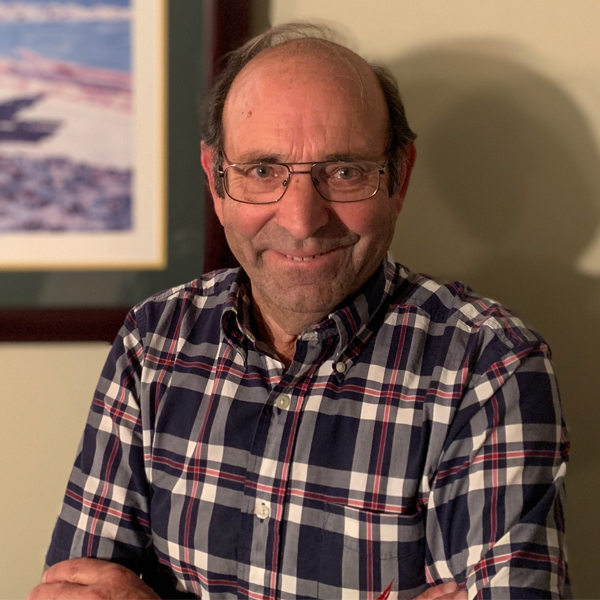 Image of Americo Silva standing up against a wall beside a picture frame wearing a plaid blue shirt