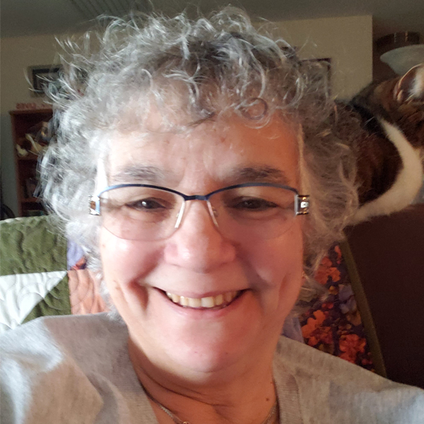 Headshot image of Hedy Halpern sitting in her chair with grey hair and glasses.