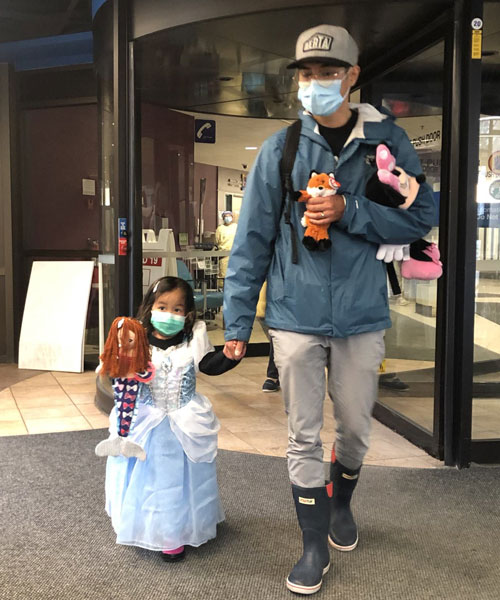 Petite fille déguisée en Cendrillon tenant la main de son père près de la porte tournante d’un hôpital.
