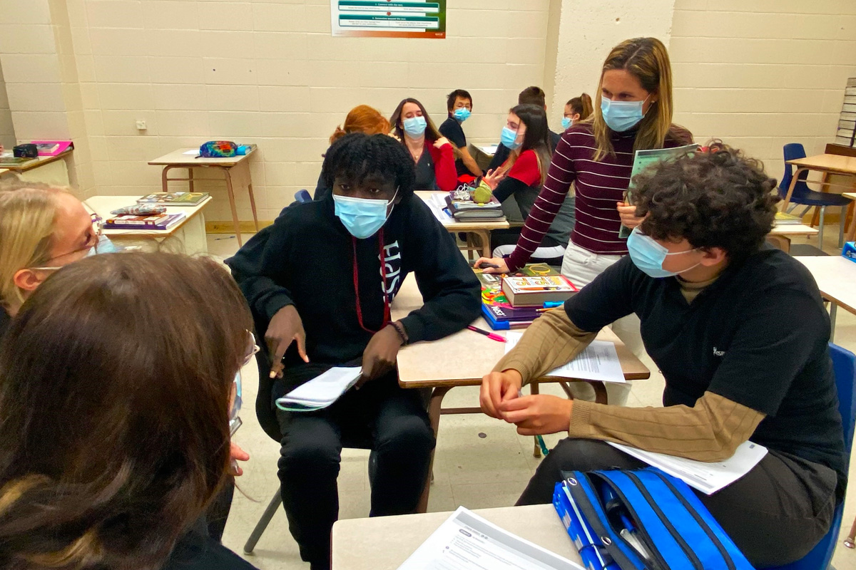 Students, wearing masks, sit in a group having a discussion while a teacher looks on