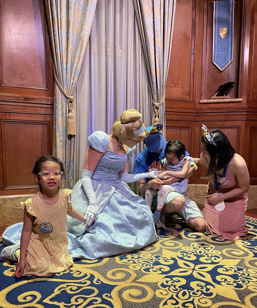 Le personnage de Cendrillon à Disney World tient la main d’un enfant bercé dans les bras de son père, tandis que la mère de l’enfant regarde et que sa sœur tient l’autre main de Cendrillon.