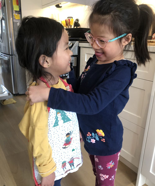 Sœur aînée avec les mains sur les épaules de sa petite sœur, toutes deux debout dans une cuisine.