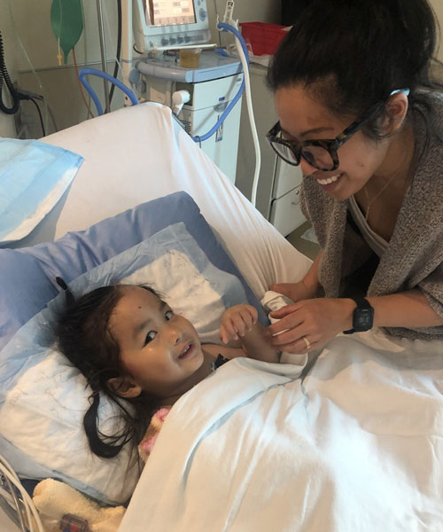 Mère debout au-dessus d’une petite fille souriante dans un lit d’hôpital.