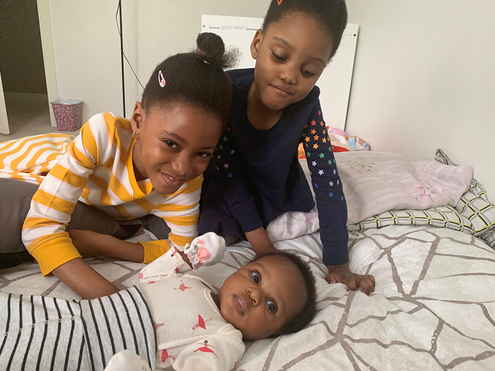 Blood recipients Aaliyah and Naila Mchopanga pose with their baby sister on a bed.
