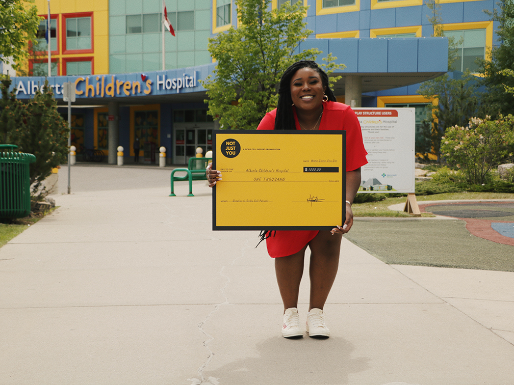 Stem cell recipient Ufuoma Muwhen holds a yellow cardboard novelty cheque in front of Calgary hospital.