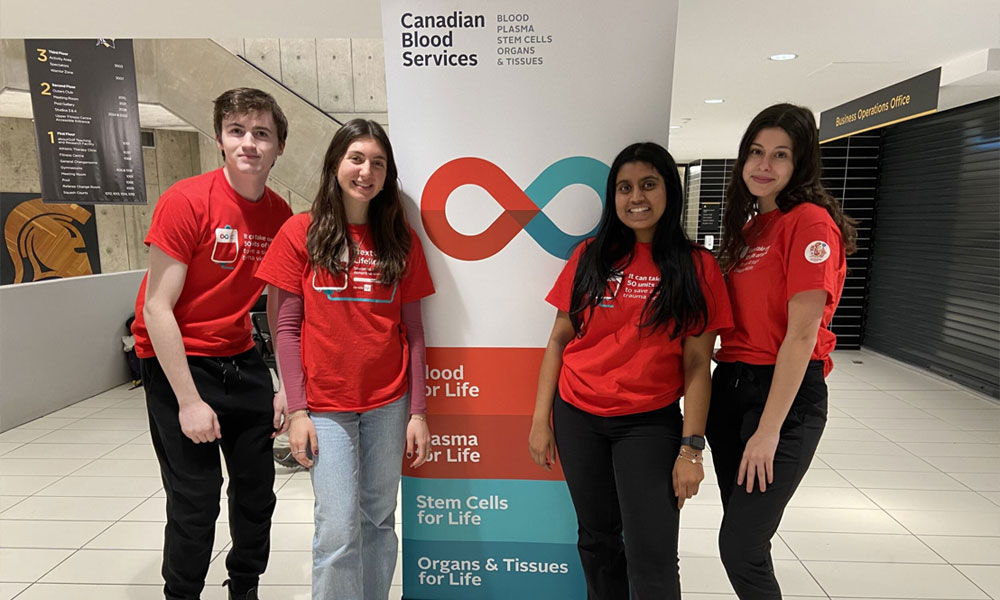 young man and three young females standing in front of a Canadian Blood Services display panel