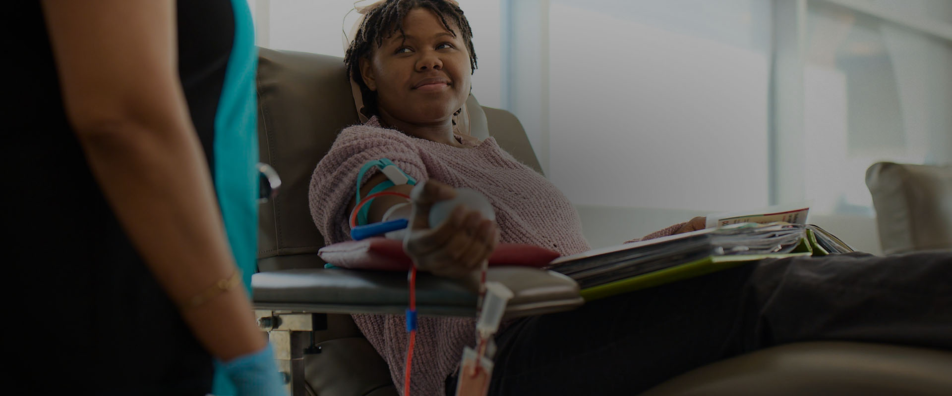 Visaree-desktop.jpg Darkly lit image of Visaree, sitting back in a donation chair while donating blood. Visaree is looking up at the nurse, while holding on to an exercise ball in the palm of her hand.