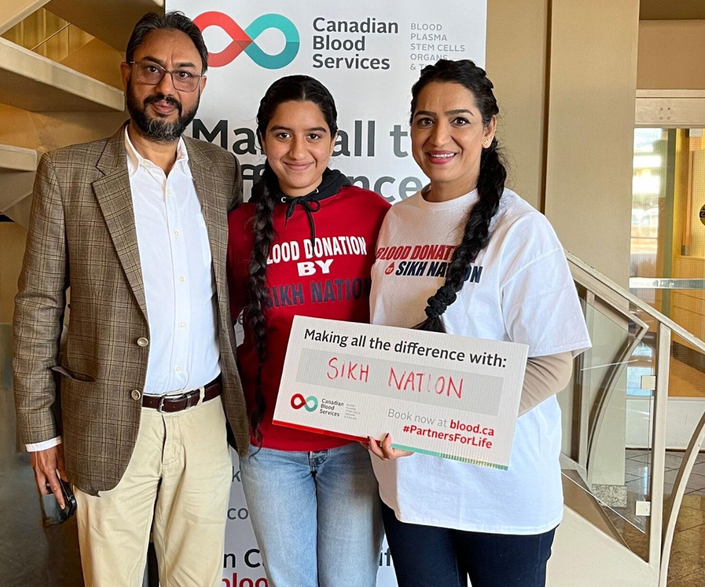 Family of three stand together holding a sign that says Making all the difference with: Sikh Nation. Two of them wear Blood donation by Sikh Nation t-shirts.