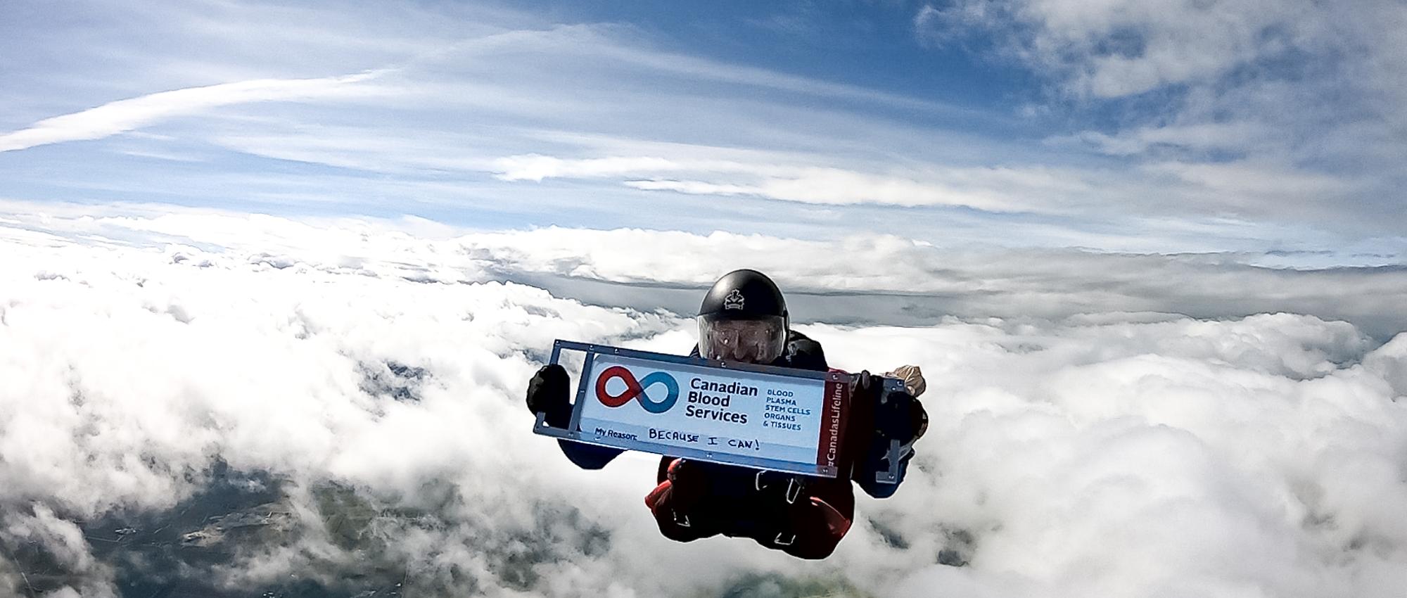 Big Image of Ian Harrop holding up a sign of Canadian Blood Services of his 600th blood donation while skydiving