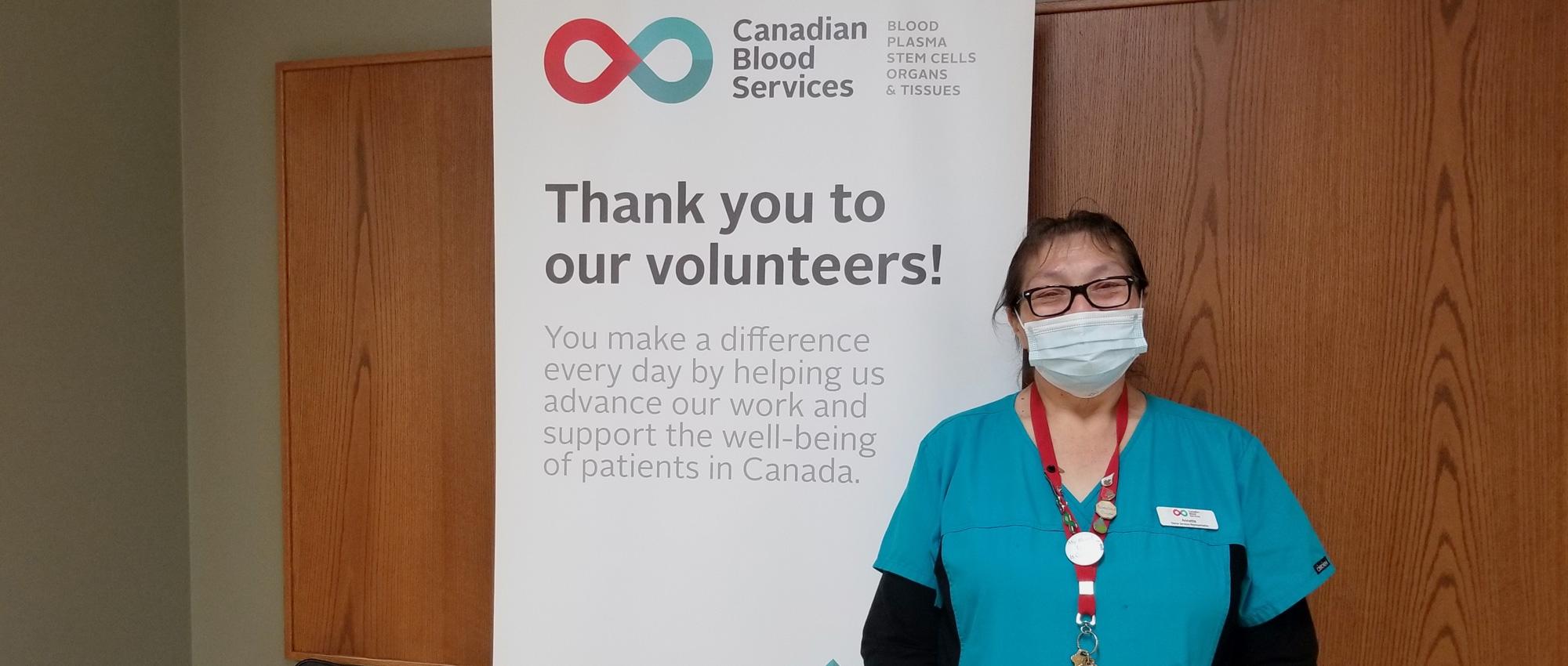 Donor services representative Annette Regalado, dressed in black and blue scrubs, stands in front of a Canadian Blood Services banner which reads: “Thank you to our volunteers.”
